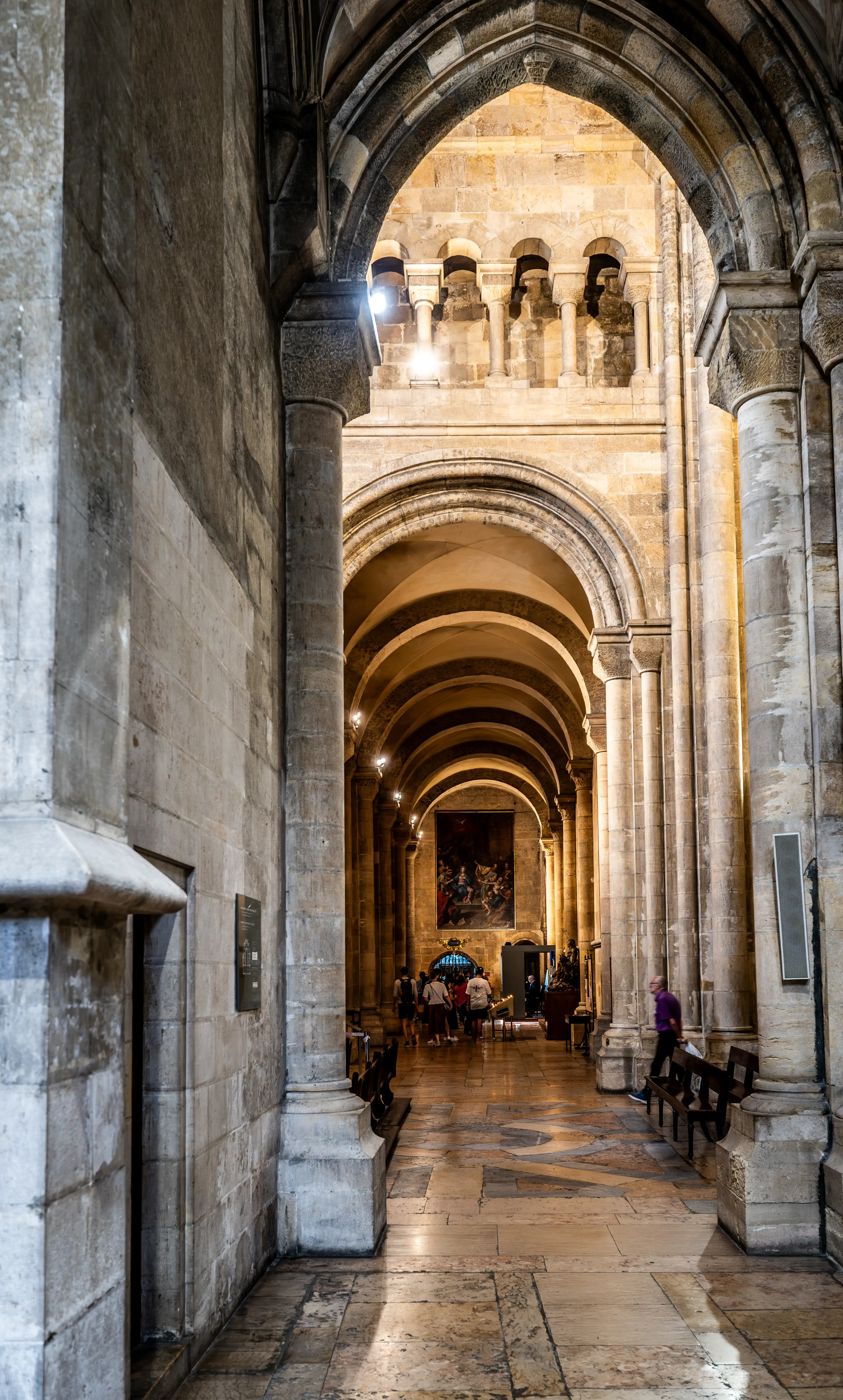 A person walks down a long, vaulted corridor with stone arches and columns inside a cathedral. Sunlight creates patterns on the floor and illuminates a large framed artwork at the far end.